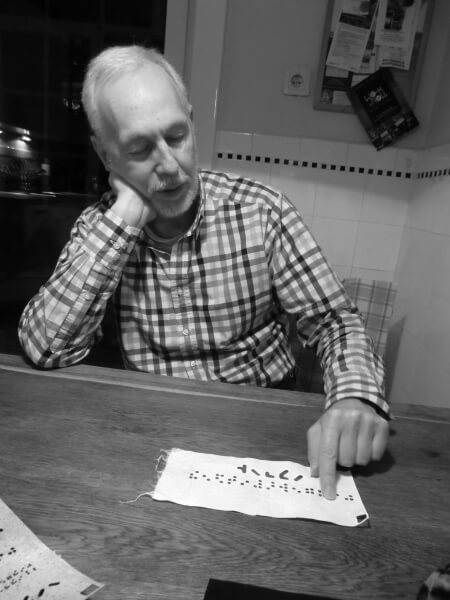 Schwarz/Weiß Foto hochkant: älterer Mann sitzt an einem Tisch und fühlt mit seinem Zeigefinger über ein Stück Stoff mit Braille-Löchern