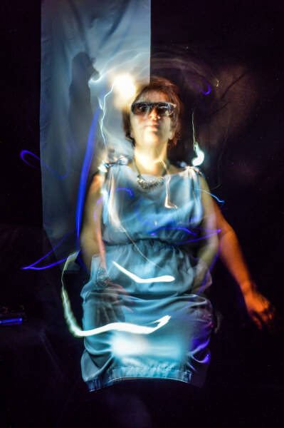 Foto hochkant, light painting, Silja Korn sitzt in einem blauen Kleid in der Mitte, es sind drei Arme von ihr zu erkennen, um sie herum Lichtstreifen, schwarzer Hintergrund