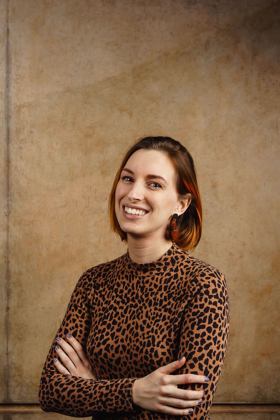 Portraitfoto Anna Flemmer, kinn-langes, braun-rotes Haar, lächelt, braun/schwarz gemustertes Rollkragen-Oberteil